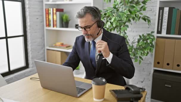 Hombre hispano joven estresado, trabajador de negocios de cabello gris, quitándose los auriculares en una oficina interior, signos de estrés laboral grave - Imágenes, Vídeo