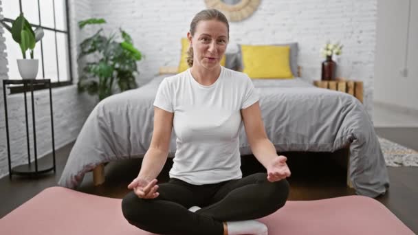 Attraktive junge blonde Frau sitzt glückselig in Yoga-Pose auf dem Bett im Schlafzimmer, spricht beim morgendlichen Videotelefonat selbstbewusst und verbessert ihre Wellness-Reise. - Filmmaterial, Video