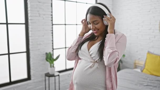 Χαμογελώντας νεαρή έγκυος στην κρεβατοκάμαρά της, βάζοντας ακουστικά στην κοιλιά της, ακούγοντας έντονα μουσική, χαλαρώνοντας στο κρεβάτι, βιώνοντας τη χαρά της μητρότητας - Πλάνα, βίντεο