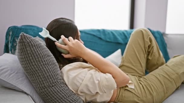 Fiatal gyönyörű spanyol nő használ okostelefon és fejhallgató feküdt kanapén otthon - Felvétel, videó
