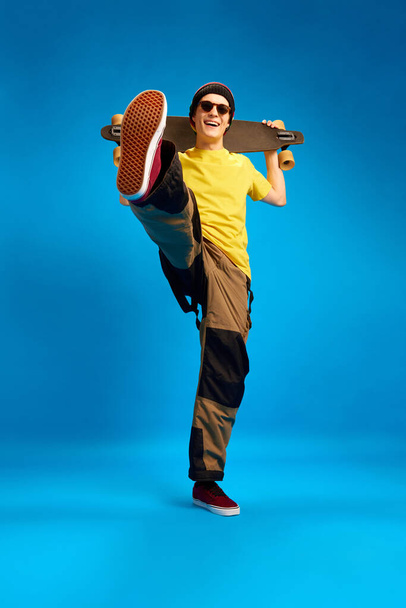 Full length portrait of cool dude rastaman, skateboarder holding skateboard and posing against blue studio background. Concept of youth, human emotions, self-expression, subcultures. - Photo, Image