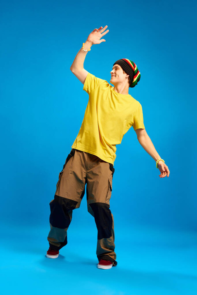 Happy, cheerful, positive young man in reggae style, rastaman cloth waves friendly against blue studio background. Concept of youth, human emotions, self-expression, subcultures, hobby. - Фото, зображення