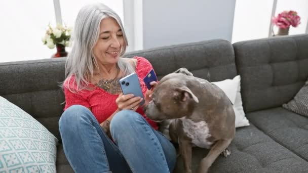 Fiduciosa di mezza età, donna dai capelli grigi gode di shopping da casa, sorridente e seduta con il suo cane su un divano, utilizzando il suo smartphone per effettuare pagamenti con carta di credito - Filmati, video