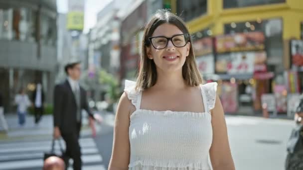 Beautiful hispanic woman in glasses, casually standing and happily smiling, posing her confidence on tokyo's modern streets, looking around, enjoying japan's urban cityscape. - Footage, Video