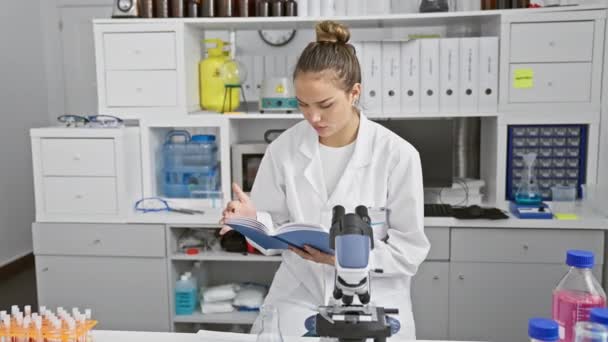 Atractiva joven científica hispana, absorta en la lectura de un libro de investigación en medio de serios trabajos de laboratorio, inclinándose sobre su microscopio en un laboratorio médico de alta tecnología - Imágenes, Vídeo