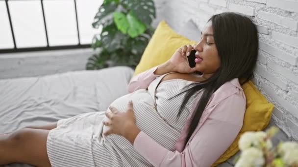 Jeune femme latine attendue, confortablement assise sur son lit, bavardant agréablement sur son smartphone tout en touchant son ventre enceinte dans le refuge réconfortant de sa chambre. - Séquence, vidéo