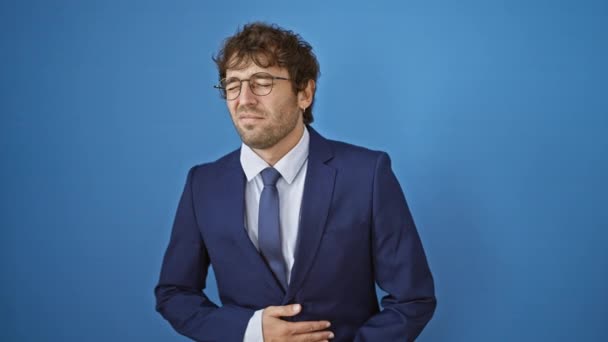 Молодий чоловік у діловому костюмі з болючим болем у животі, що зображує тугу хвороби на ізольованому синьому фоні - Кадри, відео