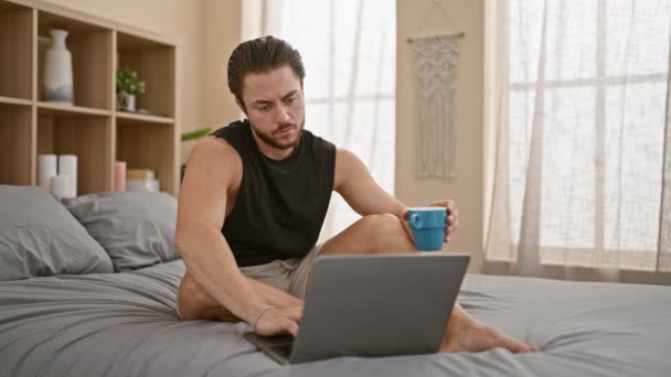 Genç İspanyol adam laptopunu kullanarak yatak odasında kahve içiyor. - Video, Çekim