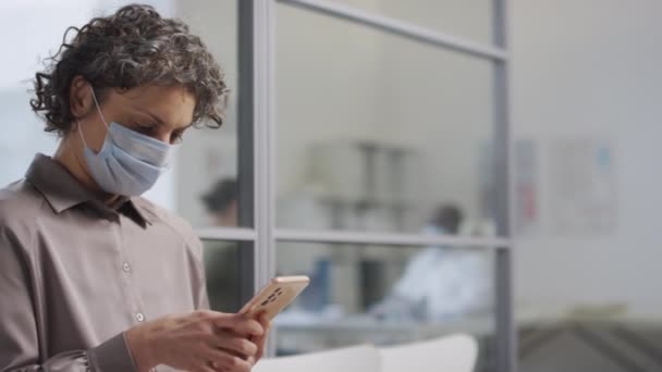 Середній постріл жінки кавказького пацієнта в масці для обличчя сидить в коридорі в клініці і чекає своєї черги під час використання мобільного телефону - Кадри, відео