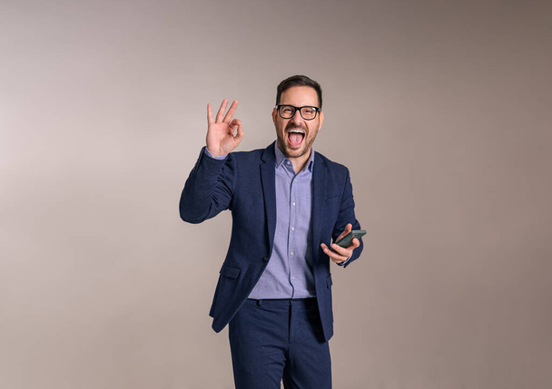 Tevreden zakenman met smartphone gebaren OK teken en schreeuwen vrolijk op witte achtergrond - Foto, afbeelding