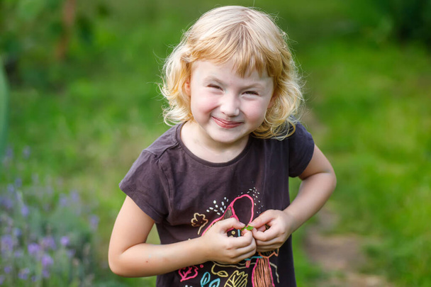 Little girl squinting her eyes while playing in the park in summer. Portrait of a cute little blond girl in the summer garden. - Photo, Image