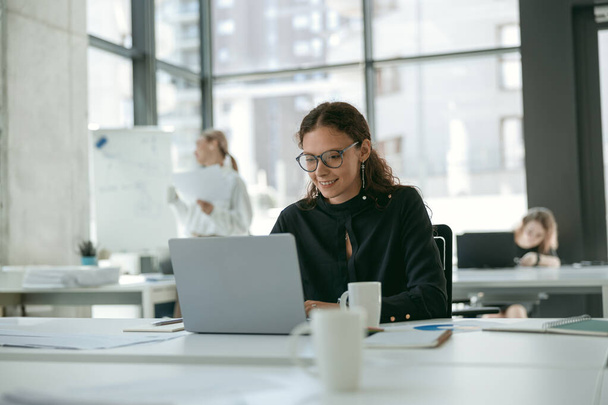Imprenditrice sorridente che lavora su laptop e beve caffè mentre è seduta in un moderno coworking - Foto, immagini