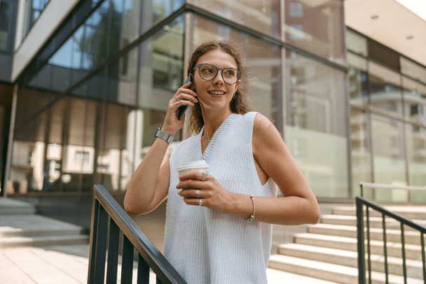 Mosolygó női szabadúszó kávéscsészével áll a modern épület háttere és beszélő telefon - Fotó, kép