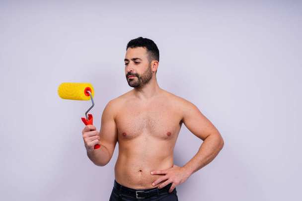 Χαρούμενος νεαρός γυμνόστηθος άντρας κρατάει ρολό μπογιάς απομονωμένο σε ένα γκρίζο στούντιο.. - Φωτογραφία, εικόνα