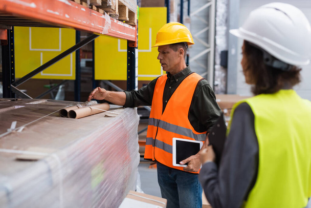 middle aged supervisor in hard hat holding tablet while explaining work to employee in warehouse - Photo, Image