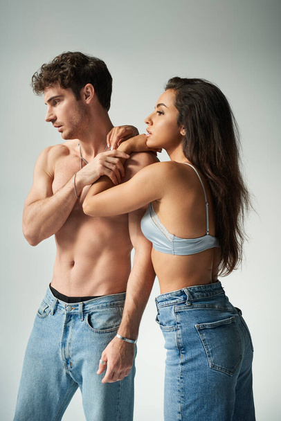 beautiful young woman in satin bra and jeans embracing shirtless man on grey background, affection - Photo, Image