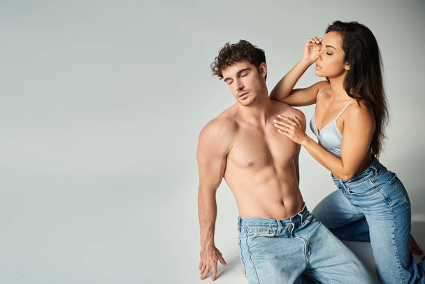 sensual young woman in silk bra and jeans posing with shirtless man on grey background, closeness - Photo, Image