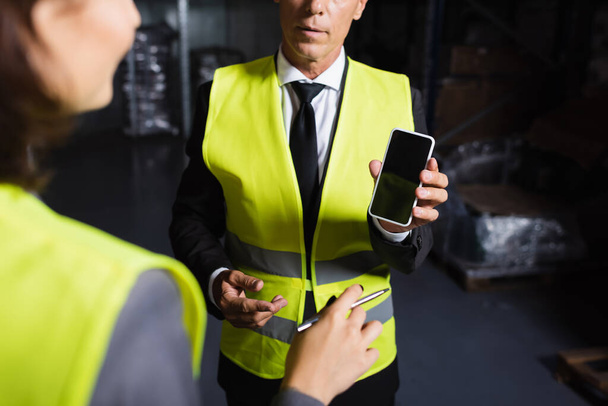 supervisor de mediana edad recortado en chaleco de seguridad usando teléfono inteligente cerca de empleada femenina borrosa - Foto, Imagen