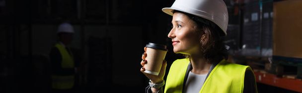 sztandar szczęśliwej kobiety w twardym kapeluszu trzymającej papierowy kubek, pracownik magazynu podczas przerwy kawowej - Zdjęcie, obraz