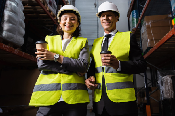 Веселая команда логистики с твердыми шляпами и кофейной улыбкой во время перерыва на складе, мужчина и женщина - Фото, изображение
