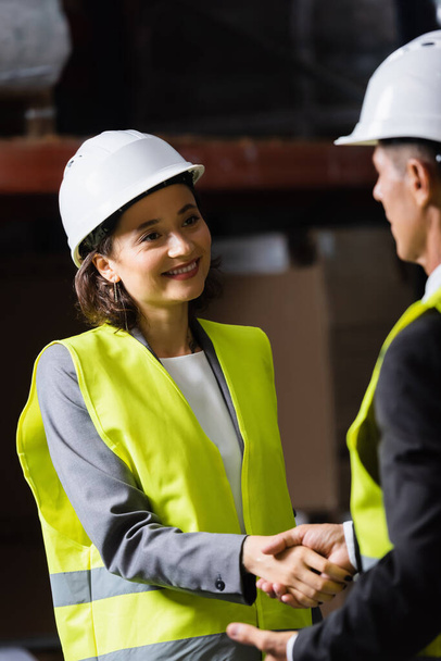 supervisor and his happy female colleague in hard hat and safety vest shaking hands in warehouse - Photo, Image