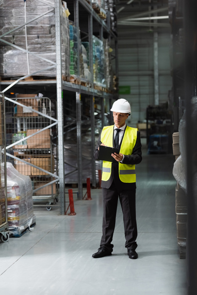 confident middle aged supervisor in safety vest and hard hat writing on clipboard in warehouse - Photo, Image