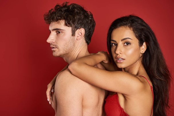 gorgeous and brunette woman in bra leaning on handsome shirtless boyfriend on red background - Photo, Image