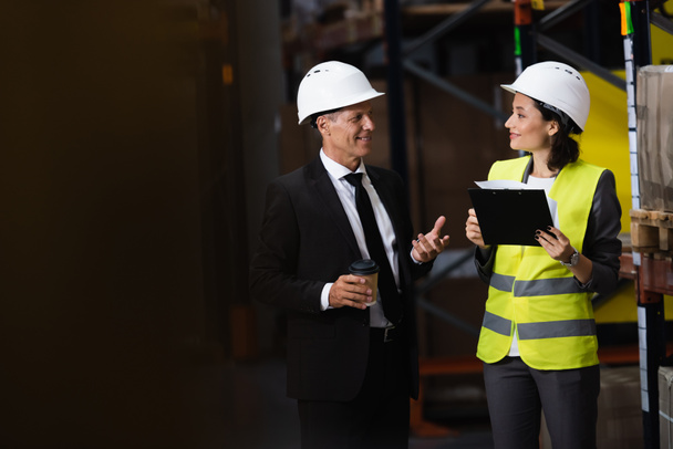 happy businessman in suit and hard hat discussing logistics operations with female employee - Photo, Image