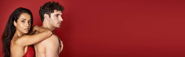 великолепная брюнетка в бюстгальтере, опирающаяся на красивого парня без рубашки на красном фоне, баннер - Фото, изображение