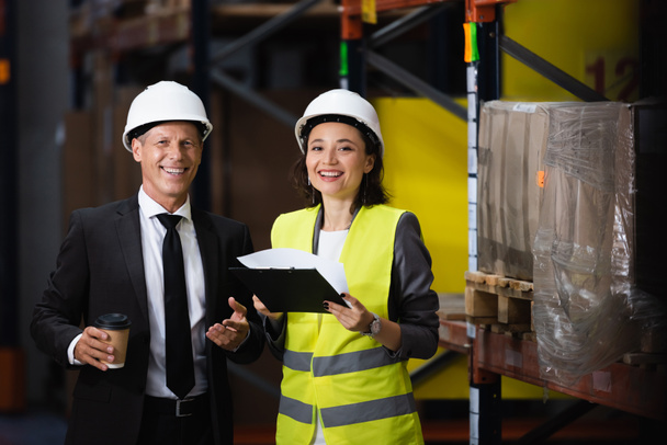 sonriente hombre de negocios en traje y sombrero duro sosteniendo café cerca de empleada femenina, concepto de logística - Foto, imagen