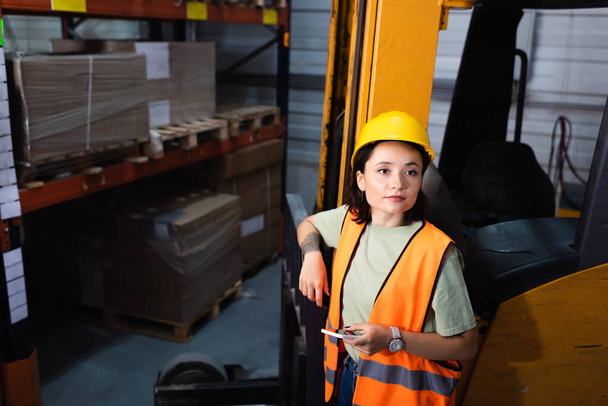 trabajadora de almacén en sombrero duro y chaleco de seguridad sosteniendo teléfono inteligente cerca de carretilla elevadora, carga - Foto, imagen