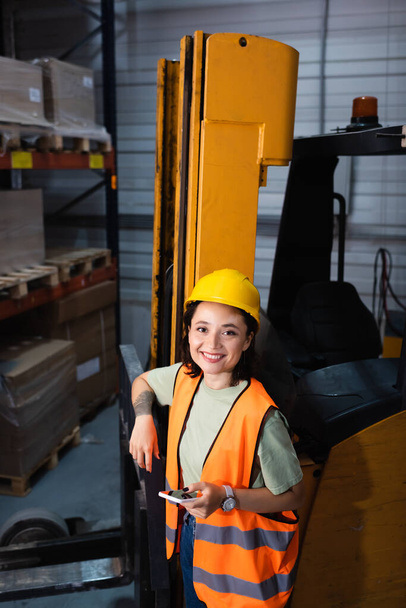 happy female warehouse worker in hard hat and safety vest holding smartphone near forklift, cargo - Photo, Image