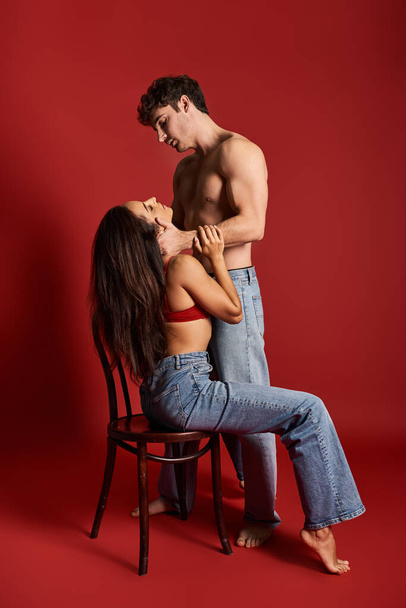 shirtless and muscular man with closed eyes embracing brunette woman on chair on red background - Foto, Imagem
