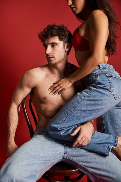 sinnliche junge Frau in Spitzen-BH sitzt auf sexy muskulösen Freund auf Stuhl, roter Hintergrund - Foto, Bild
