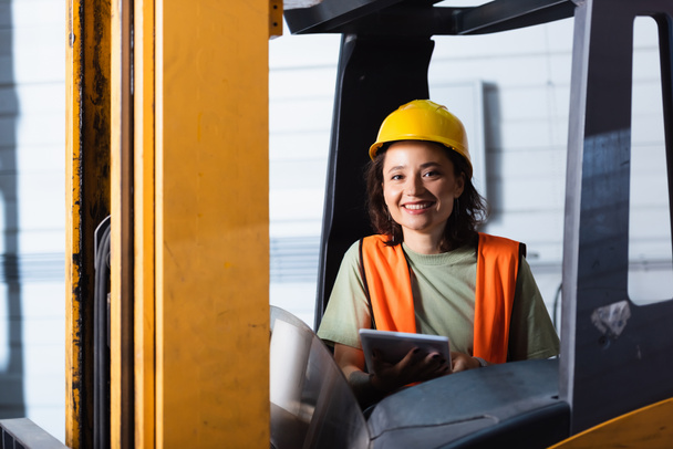 female forklift operator in hard hat and safety vest smiling and holding tablet in warehouse - Photo, Image