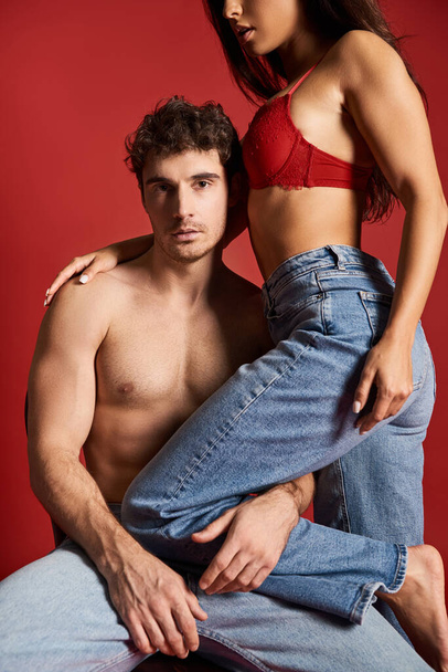 seductora mujer joven en sujetador de encaje sentado en la parte superior de sexy novio muscular en la silla, fondo rojo - Foto, imagen