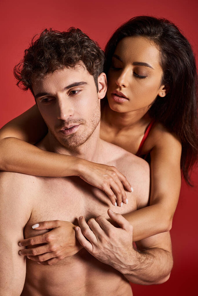 morena y hermosa mujer joven abrazando a su novio musculoso guapo sobre fondo rojo - Foto, Imagen