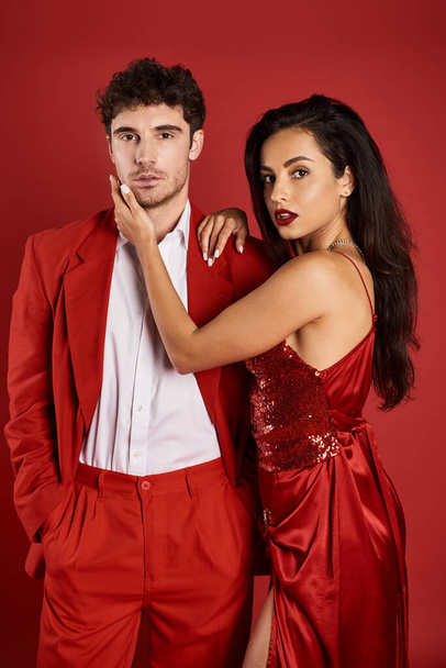 elegant woman with brunette hair embracing handsome man in formal wear on red background, glamour - Фото, зображення