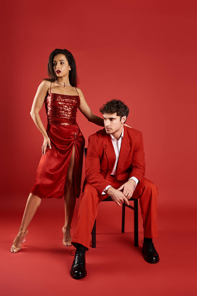 κομψή νεαρή γυναίκα σε σέξι φόρεμα στέκεται κοντά στον άνδρα σε επίσημη φθορά σε κόκκινο φόντο, εξελιγμένα - Φωτογραφία, εικόνα