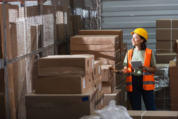 貨物の近くにデジタルタブレットを保持する安全ベストとハードハットの幸せな女性の倉庫労働者 - 写真・画像