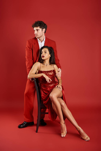 витончена молода жінка в сексуальній сукні, сидячи на стільці біля чоловіка в офіційному одязі на червоному тлі - Фото, зображення