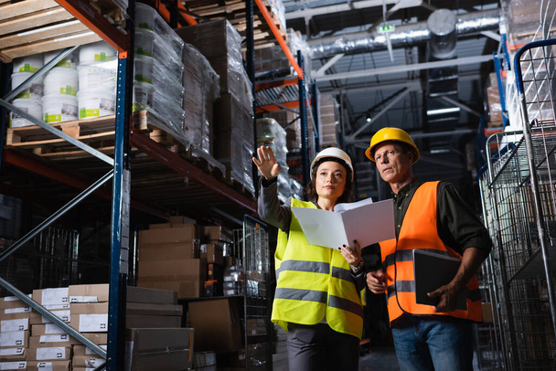 travailleurs d'entrepôt discuter de la logistique, femme avec dossier montrant la direction à un collègue d'âge moyen - Photo, image