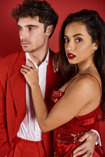 joven morena mujer con labios rojos abrazando hombre guapo en ropa formal sobre fondo rojo - Foto, Imagen