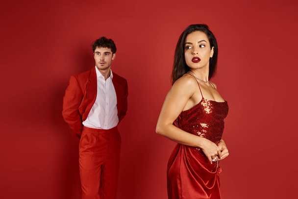összpontosítani gyönyörű és fiatal nő pózol szexi ruha közelében elmosódott férfi öltöny piros háttér - Fotó, kép