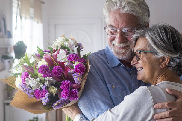 Portret van twee blije en verliefd geworden senioren of volwassen en oude mensen die thuis bloemen vasthouden en naar buiten kijken. Gepensioneerden die samen vakantie vieren en genieten. - Foto, afbeelding