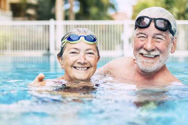ζευγάρι χαριτωμένοι ηλικιωμένοι και συνταξιούχοι στο νερό της πισίνας διασκεδάζοντας και απολαμβάνοντας μαζί - δύο ώριμοι άνθρωποι ερωτευμένοι κοιτάζοντας την κάμερα - κάνουν άσκηση και προπόνηση μαζί χαμογελώντας - Φωτογραφία, εικόνα