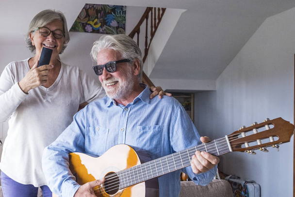 Ευτυχισμένο και αστείο ζευγάρι ηλικιωμένων και ώριμων ανθρώπων που διασκεδάζουν και απολαμβάνουν στο σπίτι κάνοντας ένα πάρτι μαζί τραγουδώντας και χορεύοντας παίζοντας κιθάρα σε εσωτερικούς χώρους. Διακοπές ή ακόμη και γιορτάζει έννοια. - Φωτογραφία, εικόνα