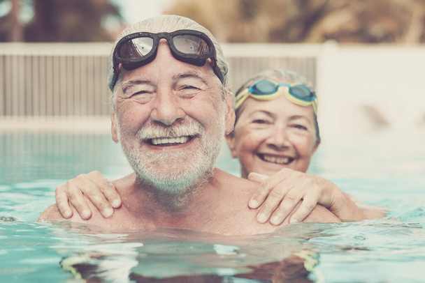 プールで2人の高齢者が一緒に抱き合って遊んでいました-幸せな成熟した人々とカメラを見ている年金受給者のカップルが笑っています - 写真・画像