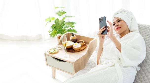 Relájese joven mujer asiática en albornoz, toalla en la cabeza, acostado en el sofá cama mientras usa teléfonos inteligentes y artículos de tratamiento de belleza para procedimientos de spa como aceite esencial y masaje con sal. - Foto, imagen