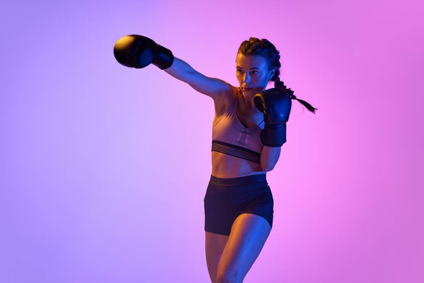 Динамичный портрет спортивной и мощной молодой женщины в боксёрской одежде, тренирующейся в движении на фоне градиентной студии в неоновом свете. Концепция спорта, активного образа жизни, здоровья, соревнований. - Фото, изображение
